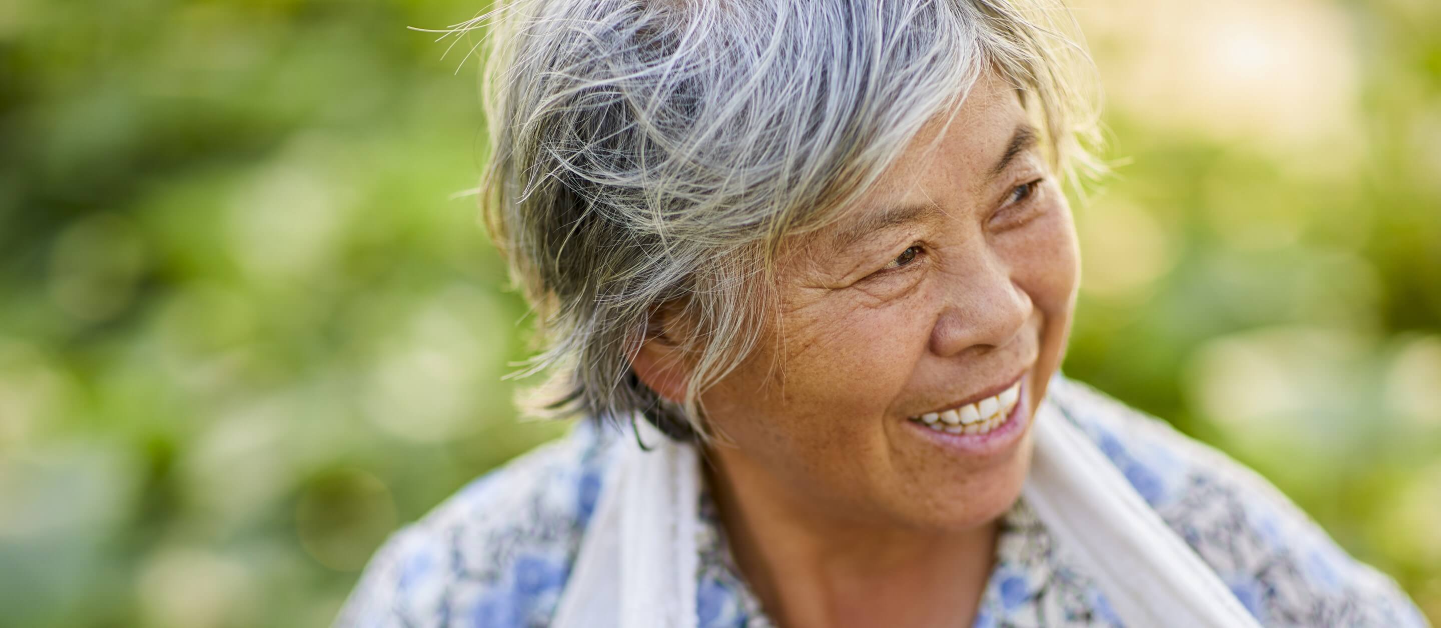 Mature Asian woman with grey hair smiling and looking to her left with bright green leaves in the background