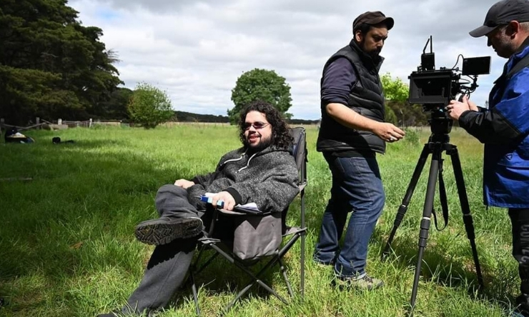 Image of young man sitting in director chair at an outdoor film set with crew members and cabers equipment in background