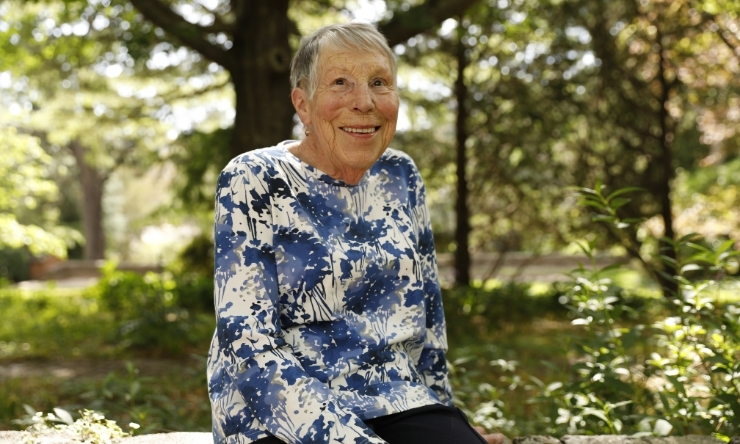 Image of smiling older woman in floral blouse sitting on garden wall with trees in background