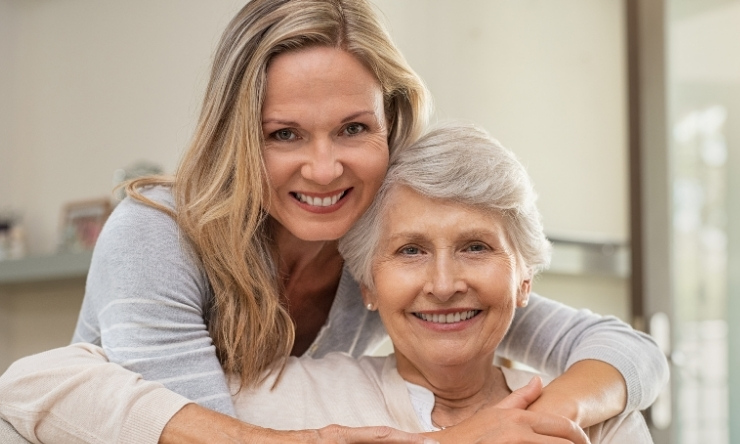 Woman in 70s sitting on couch being hugged from behind by younger woman, both happy and smiling