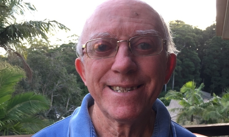 Image of smiling man with glasses and blue polo shirt with tropical trees in background