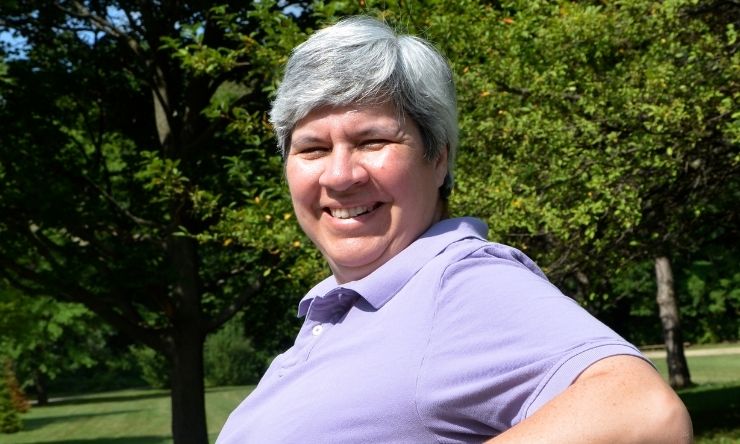 Image of woman with grey hair, wearing purple polo shirt sitting on park bench