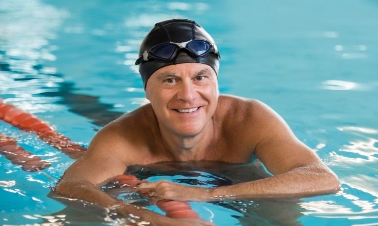 Image of man in pool leaning on edge, wearing swimming cap and goggles on head