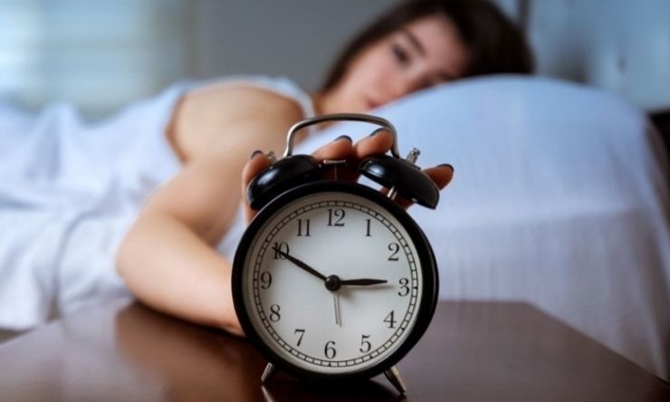 Image of an alarm clock and a woman in background lying in bed switching it off