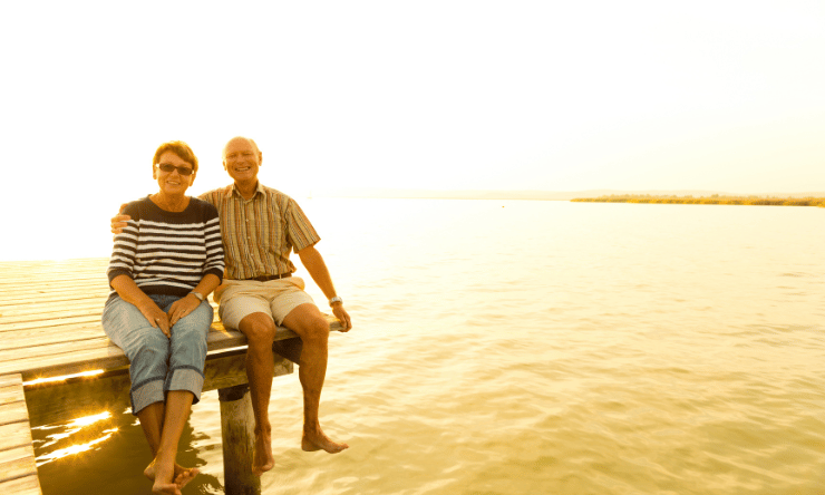 Retired couple sitting on a jetty with the sunrise behind them.