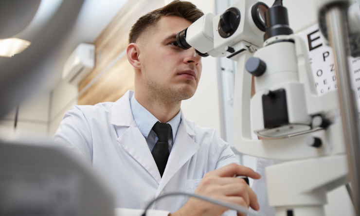 A young male optometrist wearing a white lab coat looking into the lens of a slit lamp.