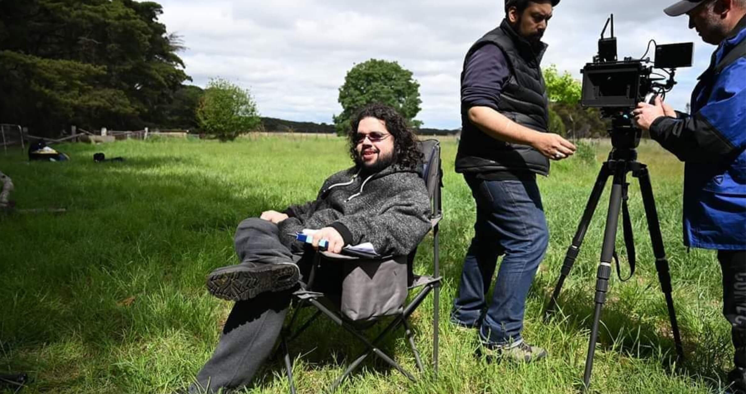 Image of young man sitting in director chair at an outdoor film set with crew members and cabers equipment in background