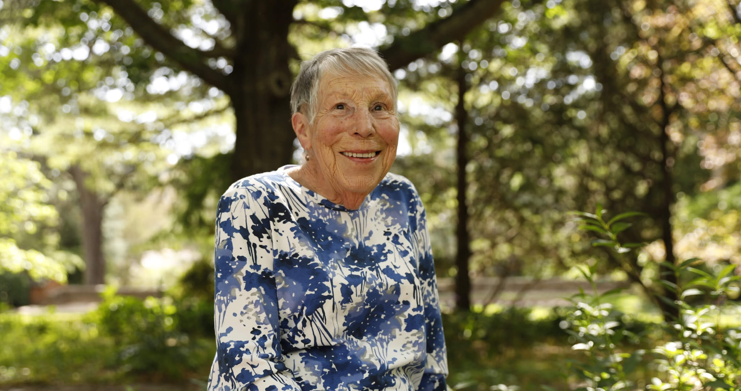 Image of smiling older woman sitting on garden wall with trees in background