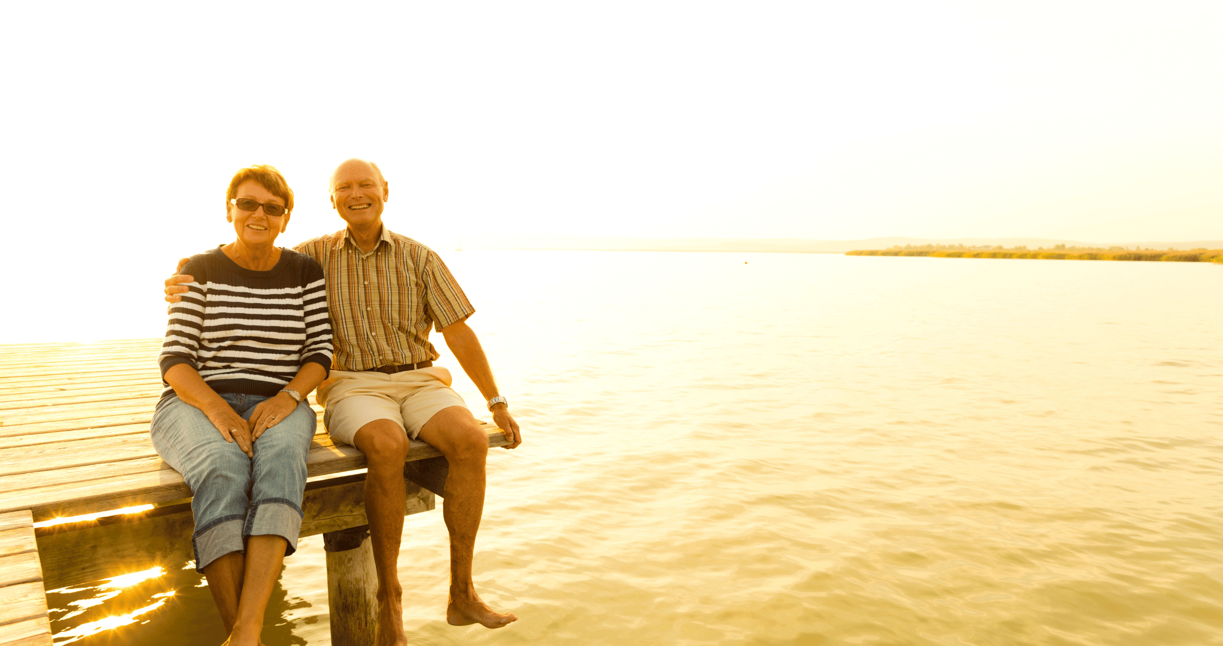 A retired couple sitting on a jetty with the sunrise behind them