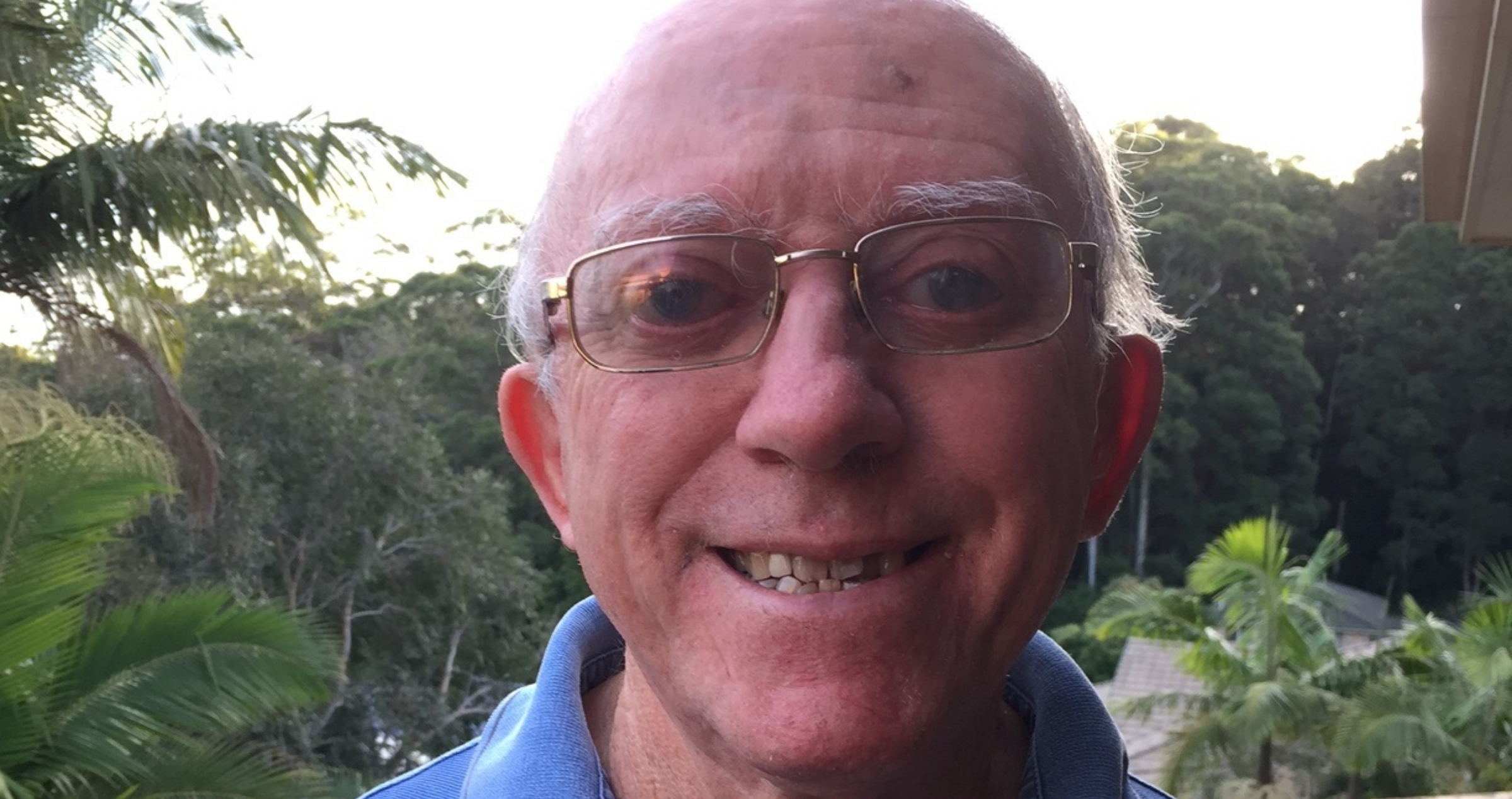 Image of smiling man with glasses and blue polo shirt with tropical trees in background