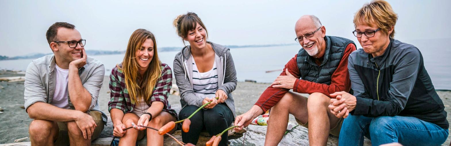 Image of family, three adult children, mother and father, sitting around fire on a beach with water in background