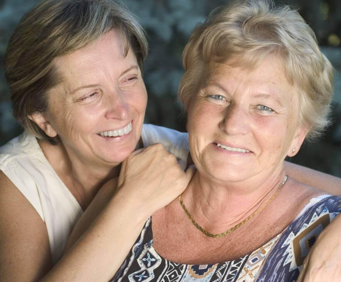 Image of two Caucasian women. Woman on the left is has her arm around the shoulder of the woman on the right and is looking at her smiling. Woman on the right is looking a the camera and smiling.