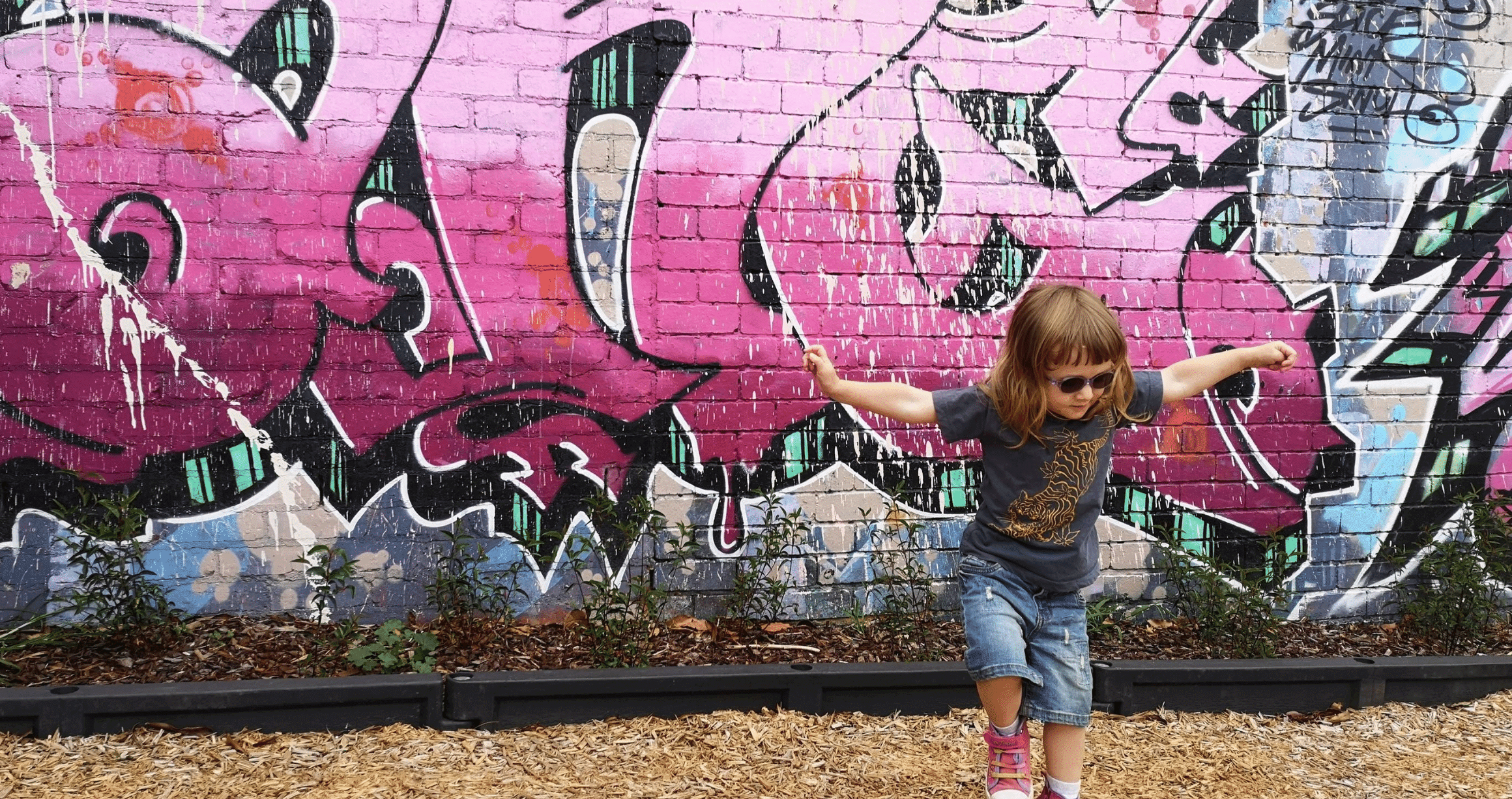 Young girl wearing glasses, playing happily in front of brick graffiti wall, very colourful