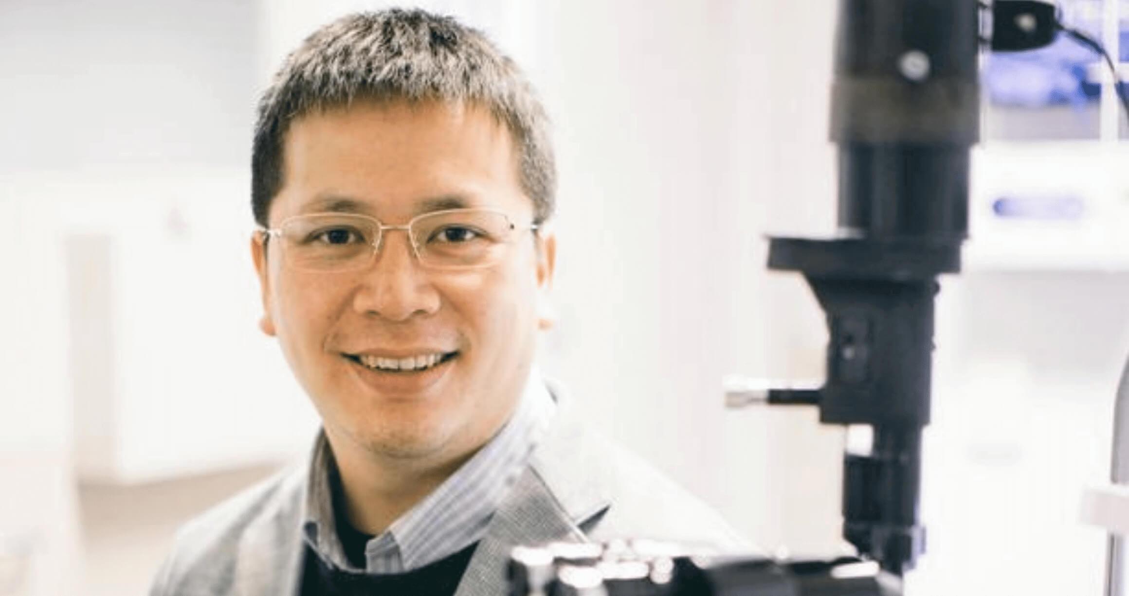 image of male doctor sitting in lab wearing coat and glasses with large microscope