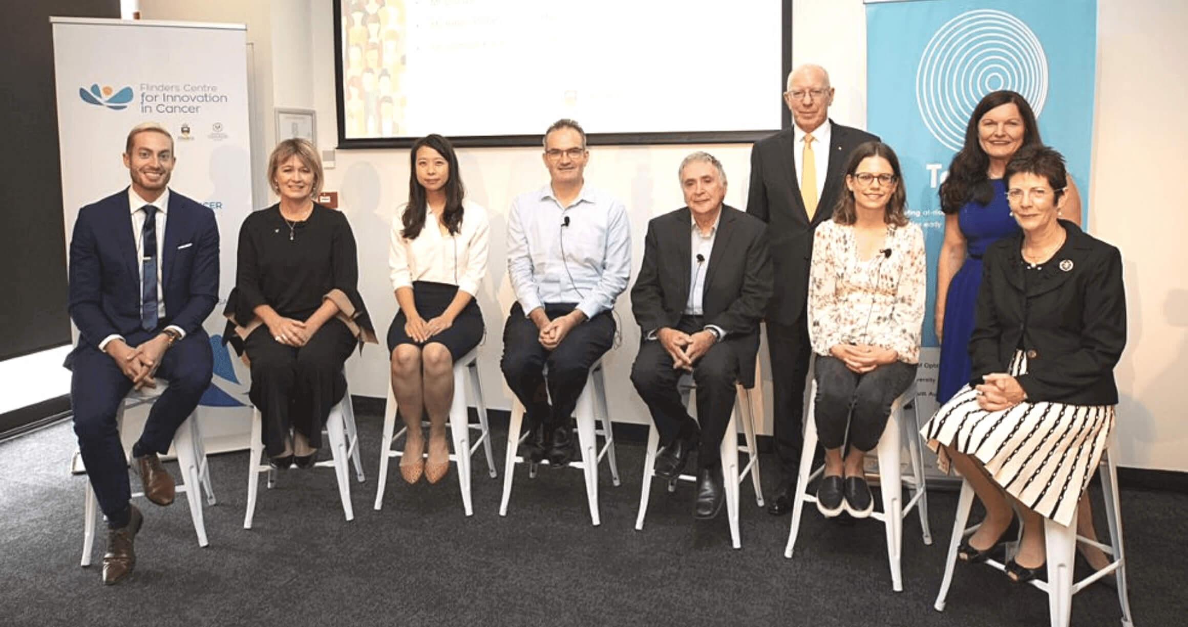 an image of seven people sitting on stools on a stage with two people standing, all smiling and with banners in the background