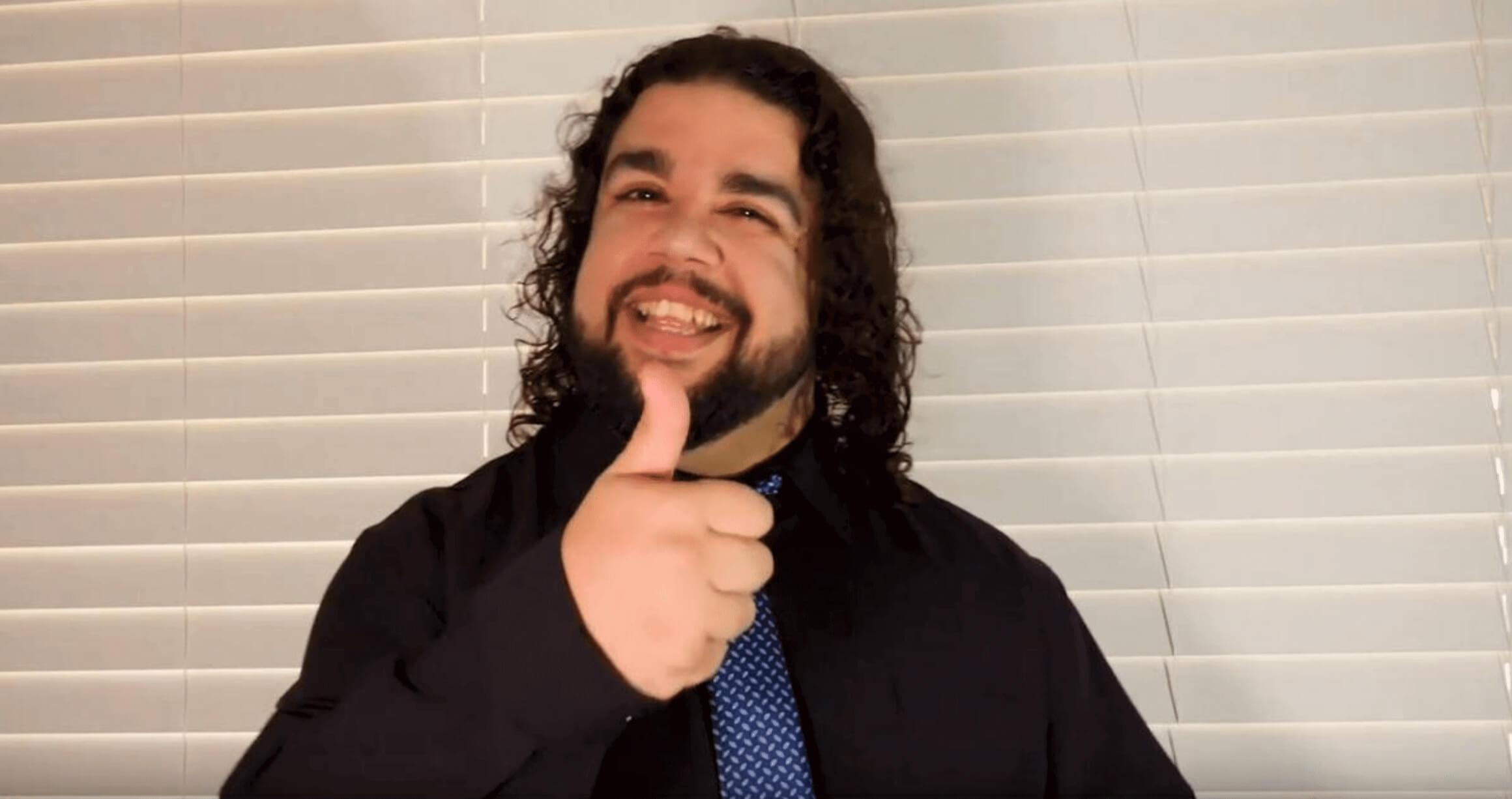 image of young man with shoulder length wavy black hair, smiling at camera and giving thumbs up