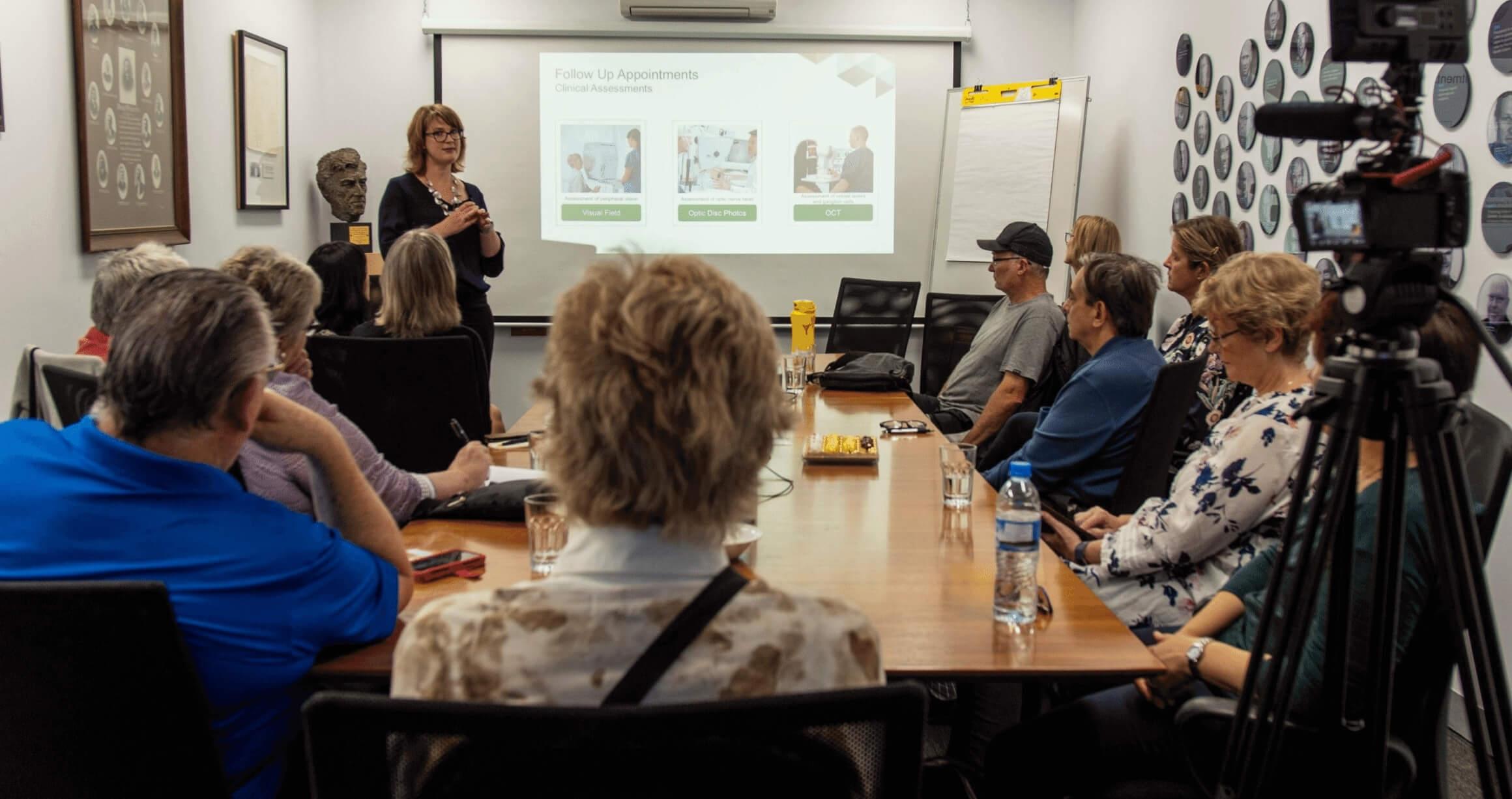 image of boardroom table with several people sitting around watching and listening to a woman give a presentation on a projector screen